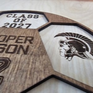May include: A laser-cut wooden sign shaped like a soccer ball, featuring the text CLASS OF 2027, the name COOPER GIBSON, and the number 22. The design includes an engraved profile of a Trojan warrior wearing a helmet with a crest.