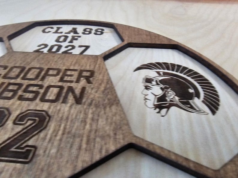 May include: A laser-cut wooden sign shaped like a soccer ball, featuring the text CLASS OF 2027, the name COOPER GIBSON, and the number 22. The design includes an engraved profile of a Trojan warrior wearing a helmet with a crest.
