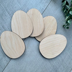 May include: Five unfinished wooden egg shapes, perfect for crafting and decorating. The eggs are arranged in a circle on a gray woodgrain surface.