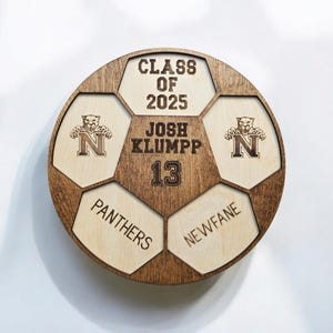 May include: A circular wooden plaque shaped like a soccer ball with dark brown stained edges and light wood panels. It features the text CLASS OF 2025, JOSH KLUMPP, 13, PANTHERS, NEWFANE, and two logos of a panther head above the letter N.