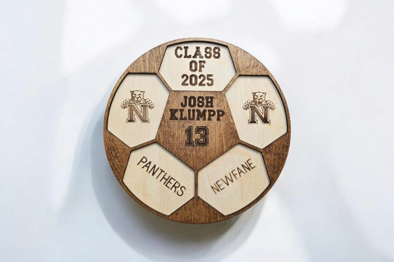May include: A circular wooden plaque shaped like a soccer ball with dark brown stained edges and light wood panels. It features the text CLASS OF 2025, JOSH KLUMPP, 13, PANTHERS, NEWFANE, and two logos of a panther head above the letter N.