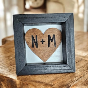 May include: A black wooden frame with a white background and a wooden heart with the letters N+M in black.