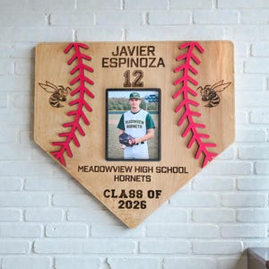 May include: A wooden baseball home plate-shaped plaque with a photo of a baseball player. The plaque has red stitching, bee graphics, and text that reads "JAVIER ESPINOZA 12", "MEADOWVIEW HIGH SCHOOL HORNETS", and "CLASS OF 2026".