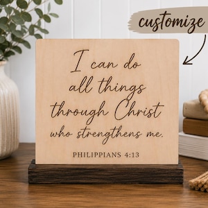 May include: A rectangular light-colored wooden sign with a dark brown wooden base. The sign features the black engraved text: I can do all things through Christ who strengthens me. PHILIPPIANS 4:13. A beige graphic bubble with the text customize it!