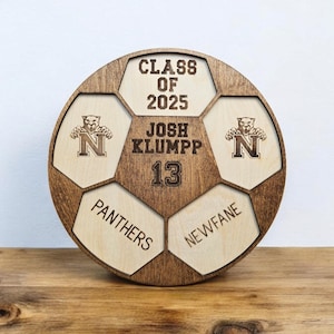 A custom engraved wooden soccer ball with the text "CLASS OF 2025"  snd player name and number on it. The ball is engraved  with a panther mascot and the words "PANTHERS" and "NEWFANE".