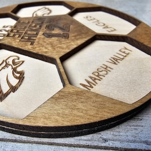 May include: A circular wooden plaque shaped like a soccer ball with laser-engraved details. It features the text SADIE JACKSON, 12, CLASS OF 2025, EAGLES, and MARSH VALLEY, along with an eagle head graphic and a hand icon within the hexagonal segments.
