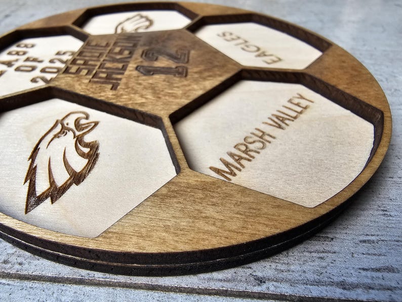 May include: A circular wooden plaque shaped like a soccer ball with laser-engraved details. It features the text SADIE JACKSON, 12, CLASS OF 2025, EAGLES, and MARSH VALLEY, along with an eagle head graphic and a hand icon within the hexagonal segments.