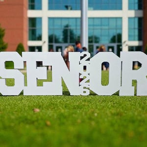 K&ouml;nnte beinhalten: Wei&szlig;e Buchstaben, die "SENIOR 2023" buchstabieren, stehen auf einer gr&uuml;nen Wiese. Die Jahreszahl 2023 befindet sich zwischen dem "N" und dem "O". Im Hintergrund sind ein Backsteingeb&auml;ude und B&auml;ume zu sehen. Die Buchstaben sind aus massivem Material.