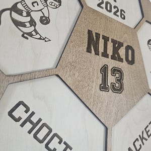 May include: A wooden wall display featuring a honeycomb pattern with engraved text and graphics. The center hexagon displays the name NIKO and the number 13. Other sections include the text CLASS OF 2026, CHOCTAW, JACKET, and a bee mascot.