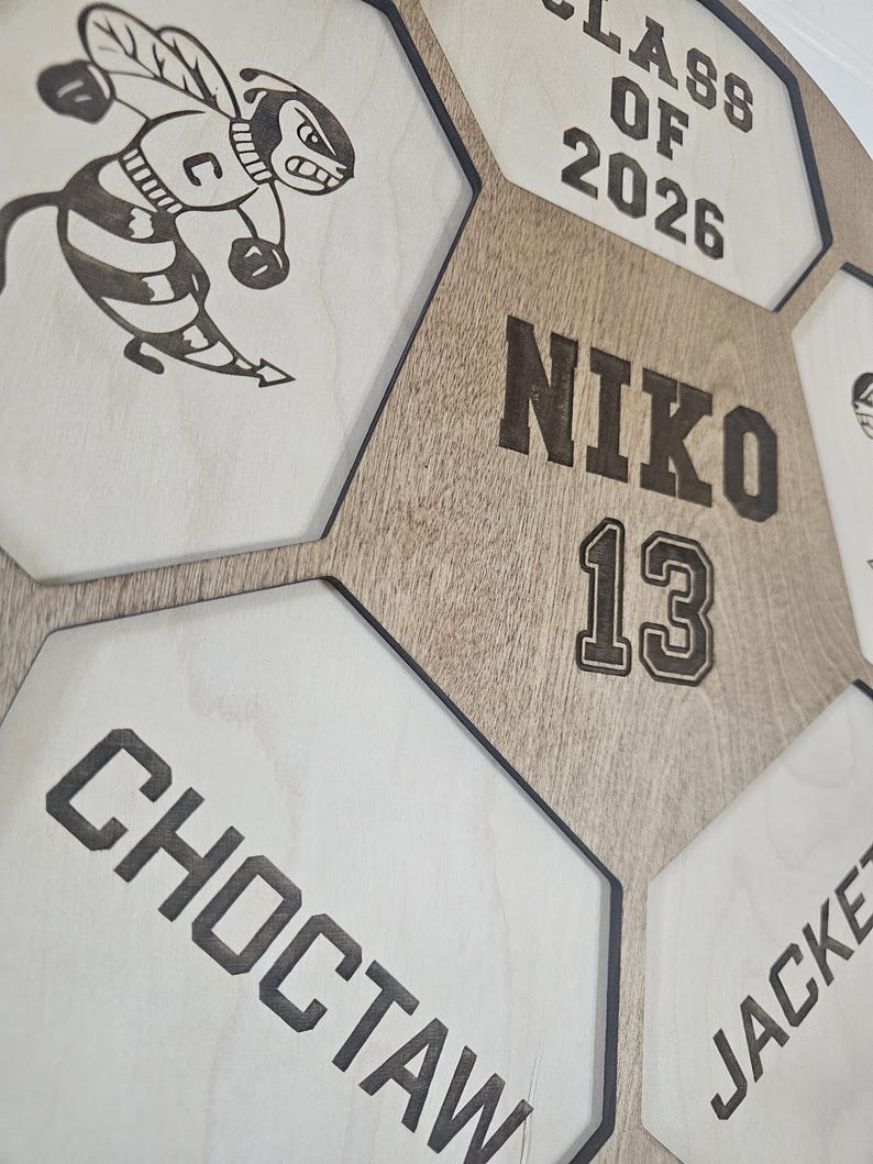May include: A wooden wall display featuring a honeycomb pattern with engraved text and graphics. The center hexagon displays the name NIKO and the number 13. Other sections include the text CLASS OF 2026, CHOCTAW, JACKET, and a bee mascot.