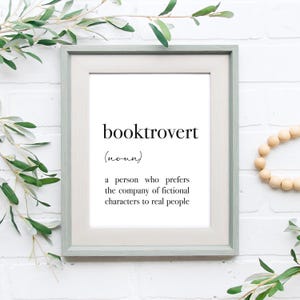 以下が含まれることがあります： 白い背景に黒い文字で「booktrovert」を定義した、額縁に入ったプリント。架空の人物との付き合いを、現実の人々よりも好む人のことを指します。