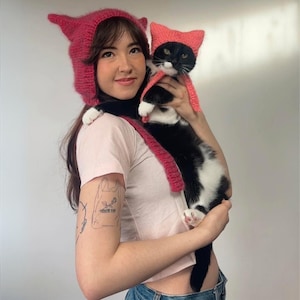 May include: A person wearing a pink knitted hat with cat ears and a pink knitted scarf is holding a black and white cat wearing a pink knitted hat with cat ears.