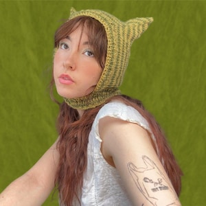 Könnte beinhalten: Eine Person trägt eine grüne und gelbe gestreifte Strickmütze mit Katzenohren. Die Mütze hat eine lange Klappe, die den Hals bedeckt. Die Person hat ein Tattoo auf dem Arm von einer Plastiktüte mit einem Gesicht.