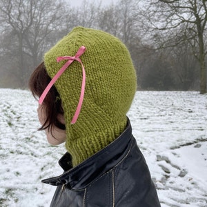 May include: A green knitted balaclava with a pink ribbon tied at the top. The balaclava is worn by a person standing in a snowy landscape.