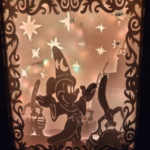 Könnte beinhalten: Eine beleuchtete Schattenbox mit einer Silhouette von Mickey Mouse, der als Zauberer verkleidet ist, einen Zauberstab hält und vor einem Schloss steht. Der Hintergrund ist ein sternenübersäter Nachthimmel.