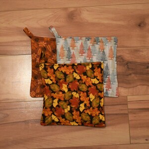 Peut inclure: Trois maniques avec des motifs sur le thème de l'automne. Une manique a un fond brun avec des feuilles orange, jaunes et vertes. Une autre manique a un fond blanc avec des arbres orange et verts. La troisième manique a un fond noir avec des feuilles orange, jaunes et vertes.