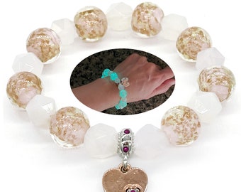 Può includere: Un braccialetto con perline bianche, rosa e dorate. Un ciondolo a forma di cuore in oro rosa con una piccola gemma rossa pende dal braccialetto. Un'immagine a incastro mostra un braccialetto luminoso al polso.