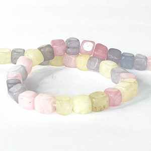 Peut inclure: Un fil de perles carr&eacute;es color&eacute;es dans les tons rose, jaune, gris et violet. Les perles sont enfil&eacute;es ensemble, formant une forme incurv&eacute;e sur un fond blanc. Les perles semblent &ecirc;tre faites d'un mat&eacute;riau lisse et poli.