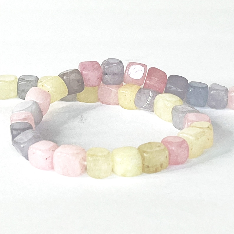Peut inclure: Un fil de perles carr&eacute;es color&eacute;es dans les tons rose, jaune, gris et violet. Les perles sont enfil&eacute;es ensemble, formant une forme incurv&eacute;e sur un fond blanc. Les perles semblent &ecirc;tre faites d'un mat&eacute;riau lisse et poli.
