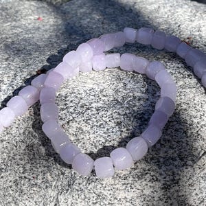 Pu&ograve; includere: Una collana di perline cubiche di colore viola chiaro semitraslucido disposte in un motivo circolare. Le perline hanno una finitura liscia e lucida e sono infilate insieme, creando un anello continuo di unit&agrave; quadrate uniformi color lavanda.