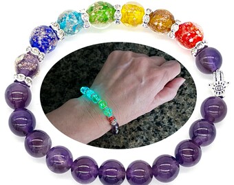 May include: A beaded bracelet featuring large, round, deep purple amethyst stones and colorful, textured beads. The bracelet includes silver-toned spacers and a Hamsa hand charm. A smaller, glowing version of the bracelet is shown in an inset.
