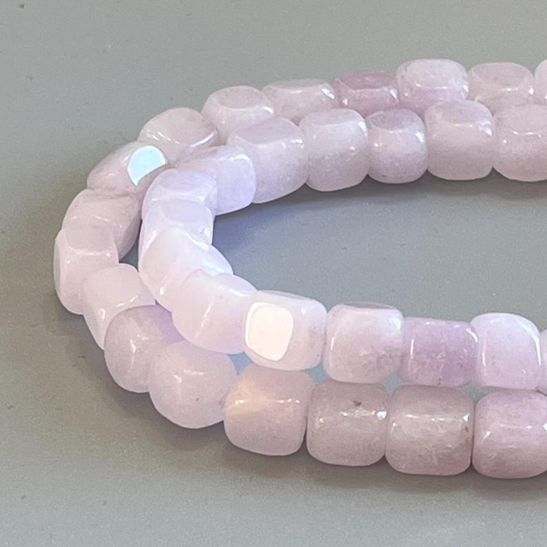 Pu&ograve; includere: Una fila di perline cubiche color viola chiaro con una finitura liscia e lucida. Le perline sono disposte in una linea curva, mettendo in risalto la loro forma quadrata uniforme e la delicata tonalit&agrave; lavanda, con riflessi di luce.