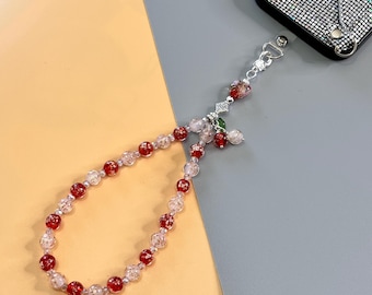 Può includere: Un ciondolo per telefono con perline rosse e trasparenti. Il ciondolo ha una chiusura argentata e un piccolo elemento decorativo con accenti rossi e verdi. La custodia del telefono è nera e ricoperta di piccoli abbellimenti lucenti.