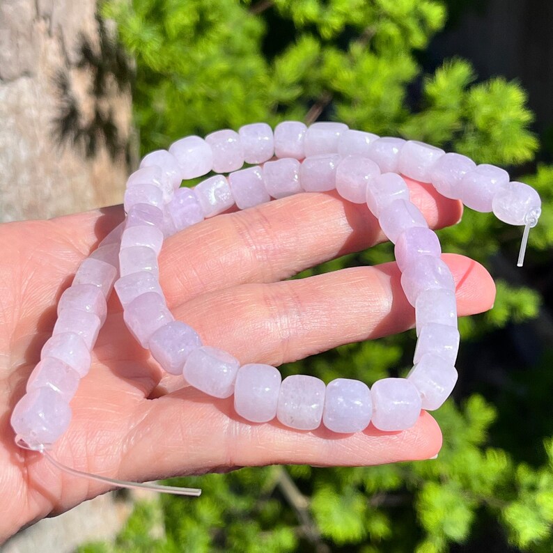 Pu&ograve; includere: Una fila di perline color lavanda chiaro a forma di cubo, infilate su un filo trasparente. Le perline hanno un aspetto liscio e semitraslucido con bordi arrotondati e una dimensione uniforme, creando una linea continua di colore viola tenue.