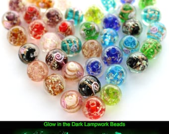 Peut inclure: Un assortiment de perles lampwork colorées et transparentes avec divers motifs internes. Les perles présentent des nuances de rouge, bleu, vert et or, certaines avec des motifs floraux ou en spirale. Le texte "Glow in the Dark Lampwork Beads" est visible.