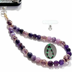 May include: A beaded phone charm in shades of purple, lavender, and clear. The charm features a variety of bead sizes and textures, with gold-colored accents. A separate white tab with a metal ring is included. A small, glowing, dark green image of the charm is also shown.