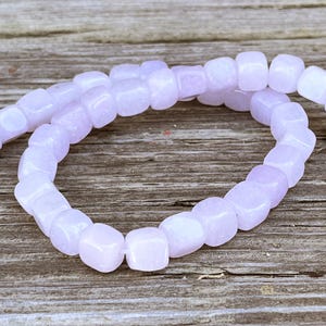 Pu&ograve; includere: Una fila di perline color viola chiaro a forma di cubo disposte in un motivo circolare. Ogni perlina presenta una superficie liscia e lucida con una tonalit&agrave; lavanda tenue e uniforme, infilate insieme per formare un anello continuo di componenti quadrati.