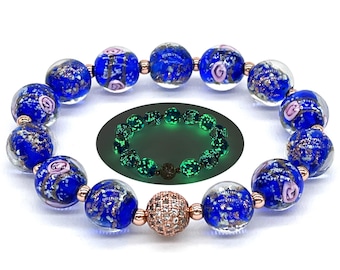 Può includere: Un braccialetto di perline con grandi perle rotonde di vetro blu con macchie dorate e vortici rosa. Il braccialetto ha dettagli metallici color oro rosa. Viene mostrato anche un braccialetto verde più piccolo e luminoso.