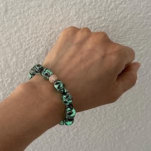 May include: A beaded bracelet featuring black and green glass beads with a floral design. The bracelet includes a rose gold-colored bead. The bracelet is worn on a wrist.
