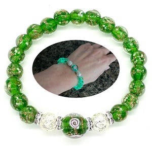 May include: A green beaded bracelet with gold flecks and silver accents. The bracelet features large, round beads with a transparent, glass-like appearance. The design includes a smaller bracelet on a wrist, showcasing the bracelet's fit and style.
