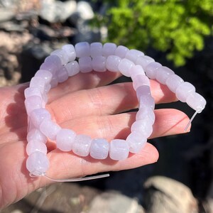 Pu&ograve; includere: Un filo di perline color lavanda chiaro a forma di cubo infilate su un filamento trasparente. Le perline hanno un aspetto liscio e semitraslucido con un colore morbido e uniforme lungo tutta la lunghezza del filo, creando un anello flessibile.