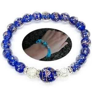 May include: Blue and silver beaded bracelet with a large central bead. The bracelet features round, translucent blue beads with gold flecks. Two silver beads flank a larger blue bead with a decorative design. A smaller inset image shows the bracelet glowing on a wrist.
