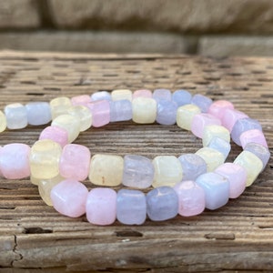 Peut inclure: Un rang de perles de pierres pr&eacute;cieuses aux couleurs pastel. Les perles sont de forme carr&eacute;e aux bords arrondis, dans des tons roses, jaunes et bleus. Les perles sont enfil&eacute;es, cr&eacute;ant un collier ou un bracelet. Le fond est une surface en bois rustique.