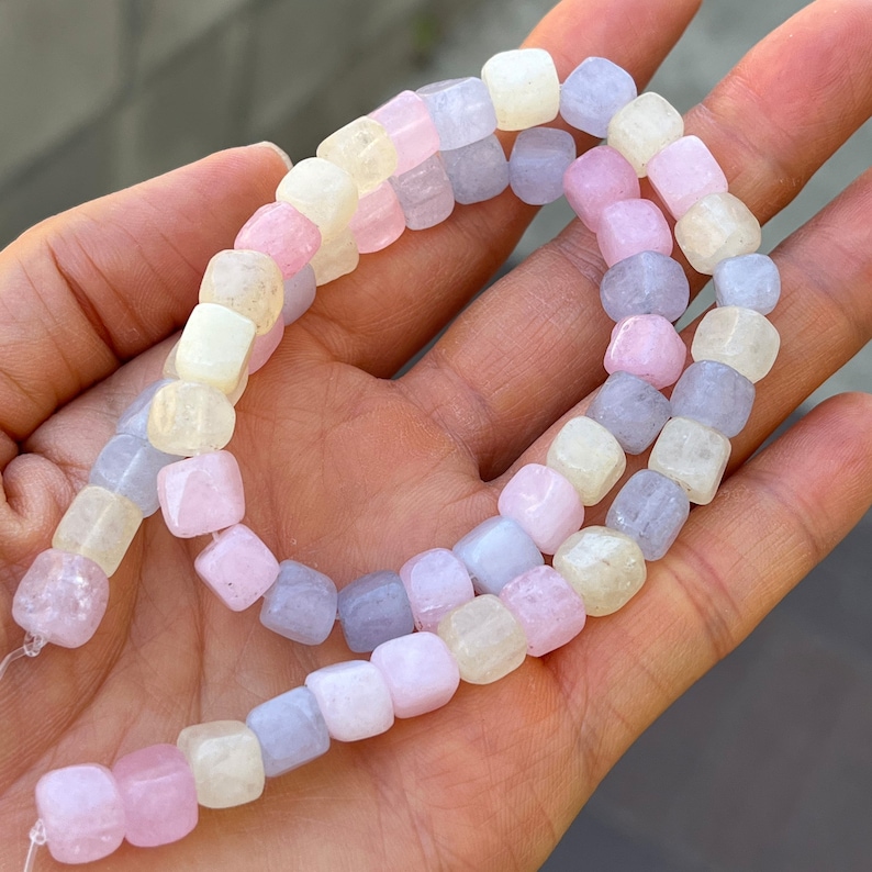 Peut inclure: Un rang de perles carr&eacute;es de couleur pastel dans les tons rose, lavande et cr&egrave;me. Les perles sont enfil&eacute;es ensemble, cr&eacute;ant un collier ou un bracelet. Les perles sont tenues dans une main.