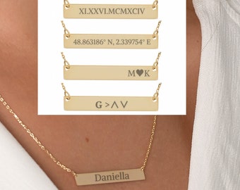 May include: A gold chain necklace with five bar pendants. The pendants are engraved with the text "XI.XXXVI.MCMXCIV", "48.863186° N, 2.339754° E", "M♥K", "G>AV", and "Daniella".