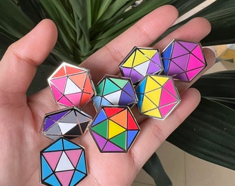 May include: A collection of seven metal enamel pins shaped like twenty-sided dice. Each pin features a unique geometric pattern with vibrant color combinations including pink, blue, yellow, orange, purple, green, and white, all outlined in silver.