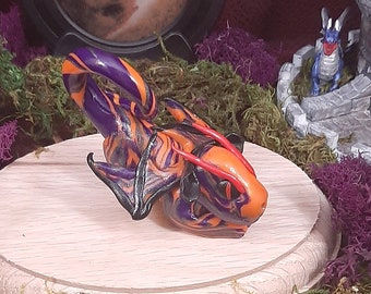 May include: A small, hand-painted glass dragon figurine with orange, black and purple colours. The dragon is perched on a wooden base and surrounded by green moss.