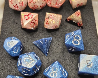 May include: A selection of polyhedral dice in different shapes. Some are translucent pink with red numbering and glitter, others are a sparkling blue with silver numbering. These dice are for tabletop gaming.