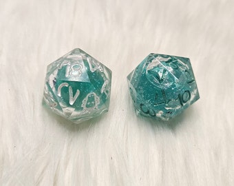 May include: Two clear, teal-coloured dice with white numbers. The dice are on a white, fluffy surface.