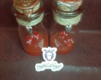 May include: Two glass potion bottles filled with red liquid, sealed with corks and twine. The corks have brown wax seals. Inside each bottle is a red, multi-sided die. A sticker with a goat head and the text "mythical myst" is at the bottom.