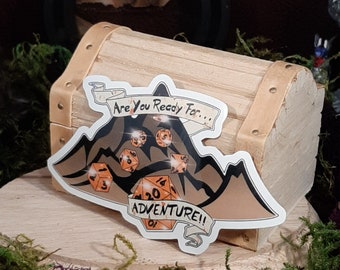 May include: A white sticker featuring a mountain range design with the text "Are you ready for... Adventure!" The mountain range is made of black lines and has orange dice scattered throughout.