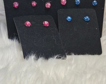 May include: Several pairs of stud earrings shaped like miniature twenty-sided dice. The earrings feature glittery pink and blue translucent resin with white or black numbering, each mounted on individual black rectangular display cards.