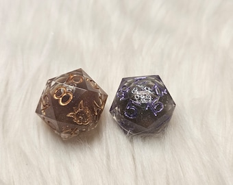 May include: Two clear dice with gold and purple glitter embedded in the resin. The dice are a dark brown and a dark purple colour. The dice are on a white fluffy surface.