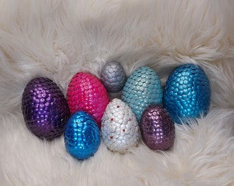 May include: A collection of eight egg-shaped objects covered in overlapping metallic scales. The eggs feature various colours including purple, hot pink, silver, light blue, bright blue, and white with small red accents embedded in the white scales.