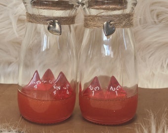 May include: Two clear glass bottles with cork stoppers and heart charms. Each bottle contains red liquid and red dice. The bottles are decorated with twine and sit on a brown surface against a white, fluffy background.