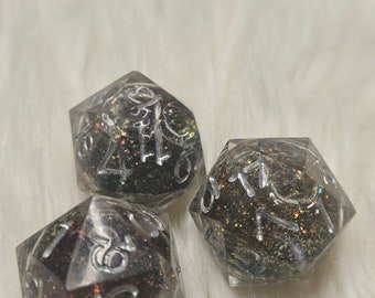 May include: Three clear, iridescent dice with black and silver glitter embedded in the resin. The dice are numbered 1-6 and have a black background with silver numbers.